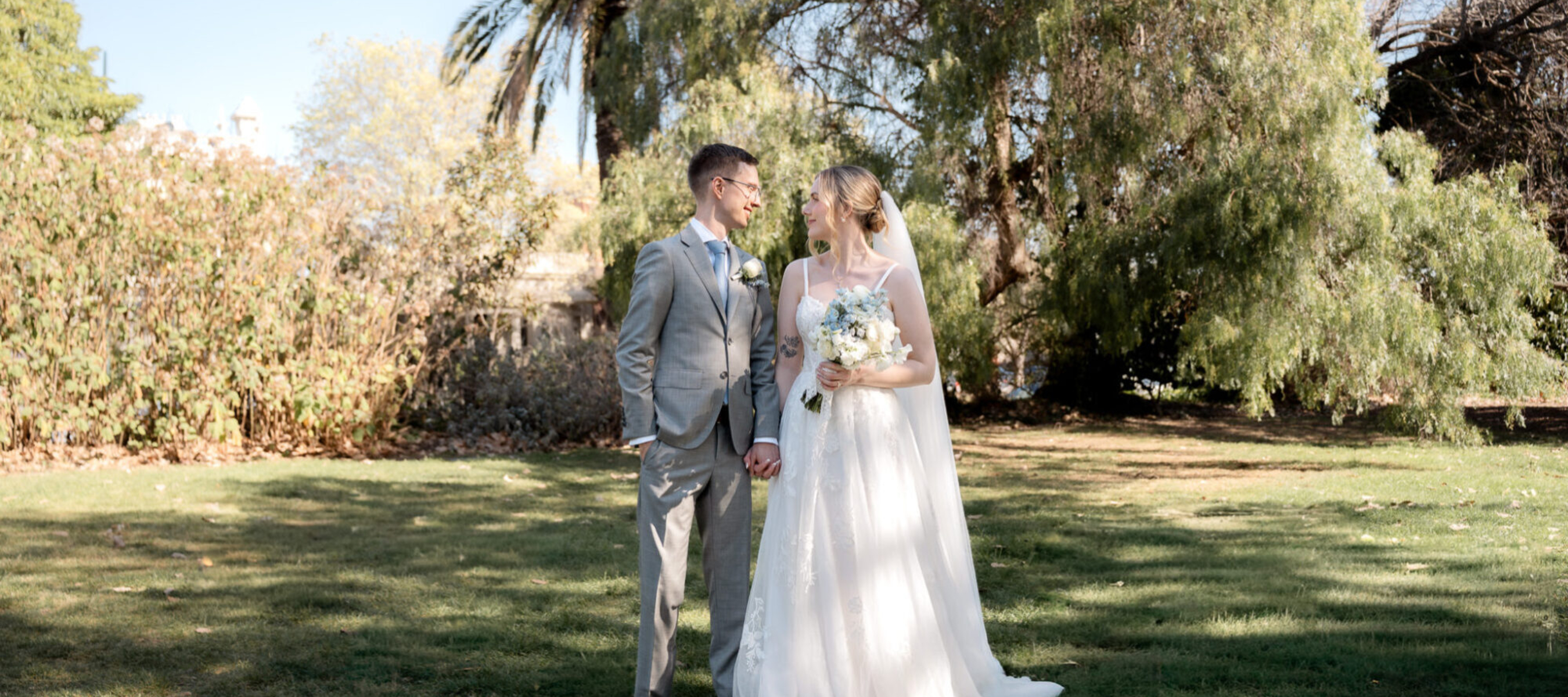 Bride and Groom standing side by side holding hands looking at eachother. Surrounded by trees and open grass area.