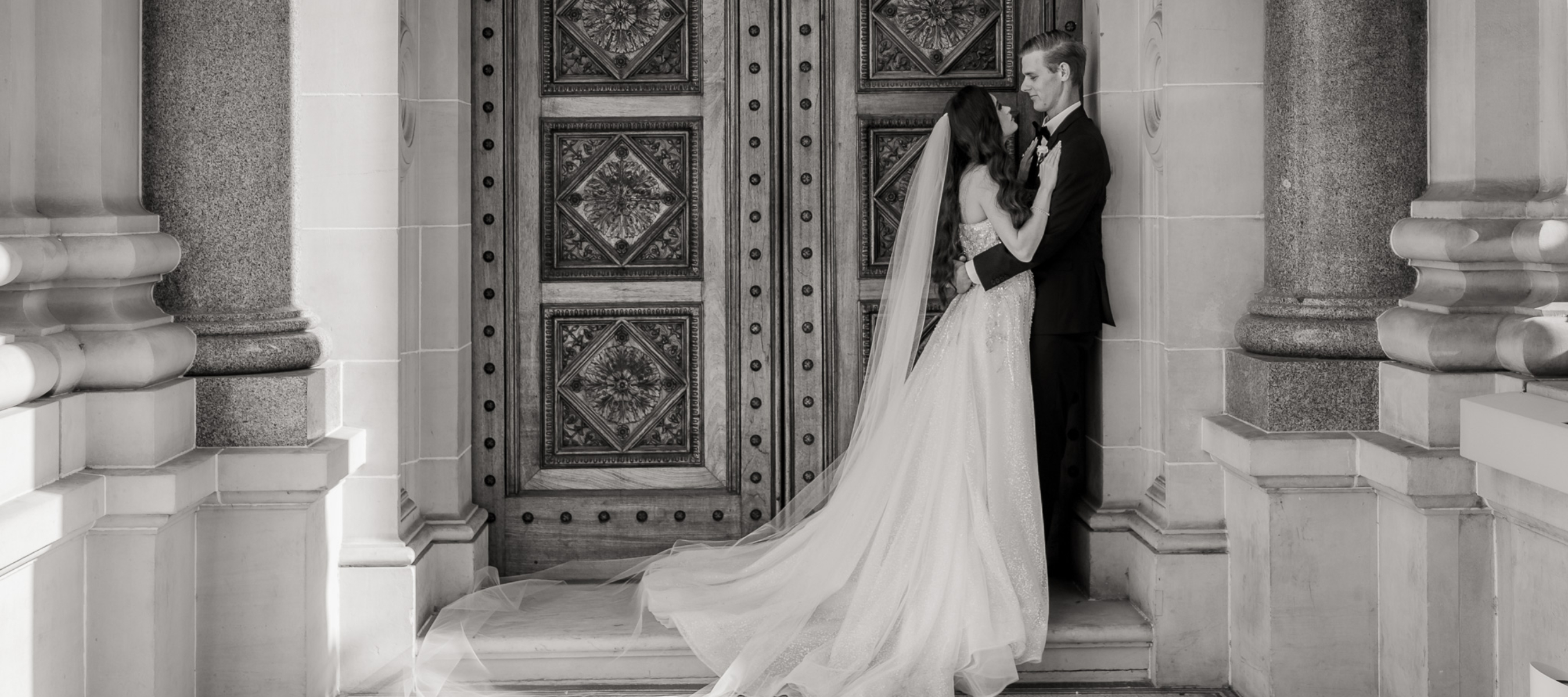 Bride looking and leaning on her Groom who is leaning against a wall in front of an old engraved door. Stone pillers around and tiled floors are surrounding them.
