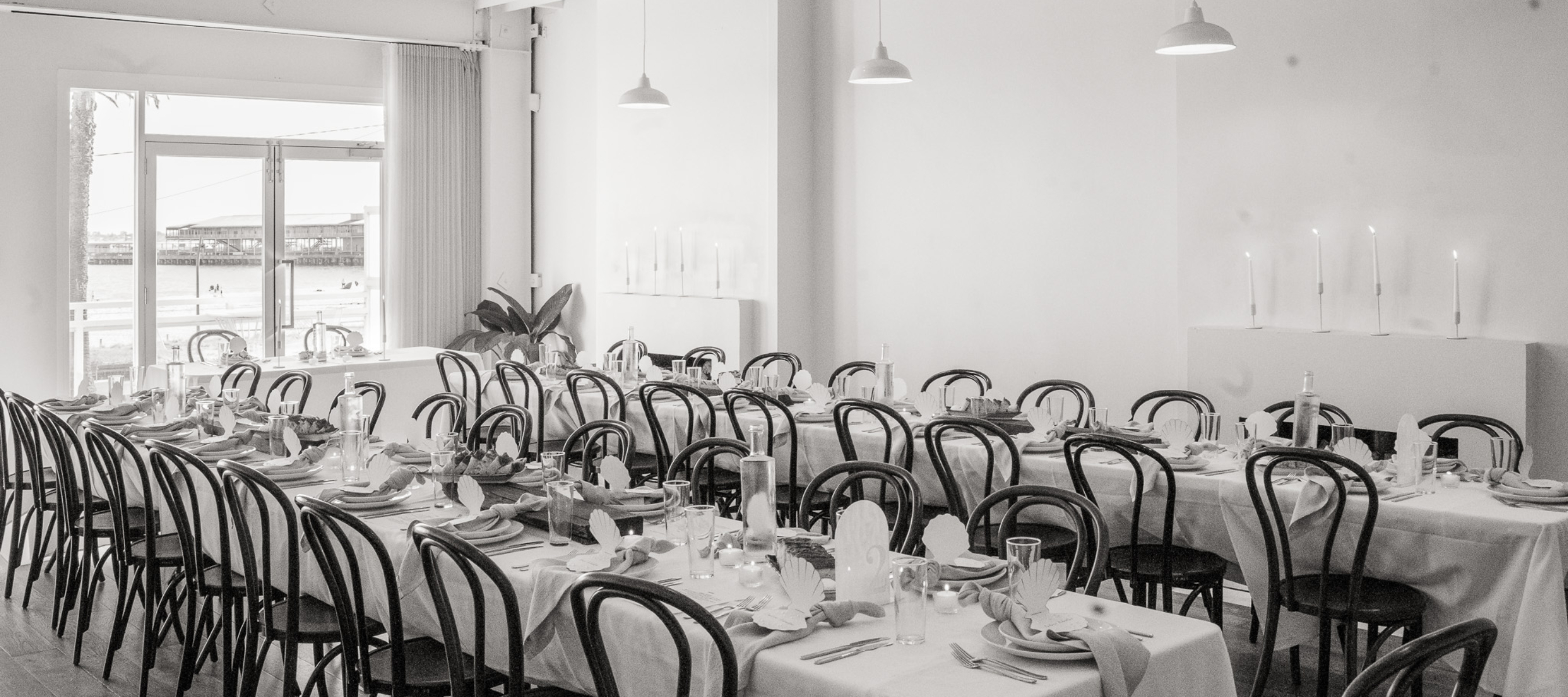 Black and white image of a wedding venue reception space. Long tables dressed in white table clothes with chairs, candles, fireplace and table settings. .