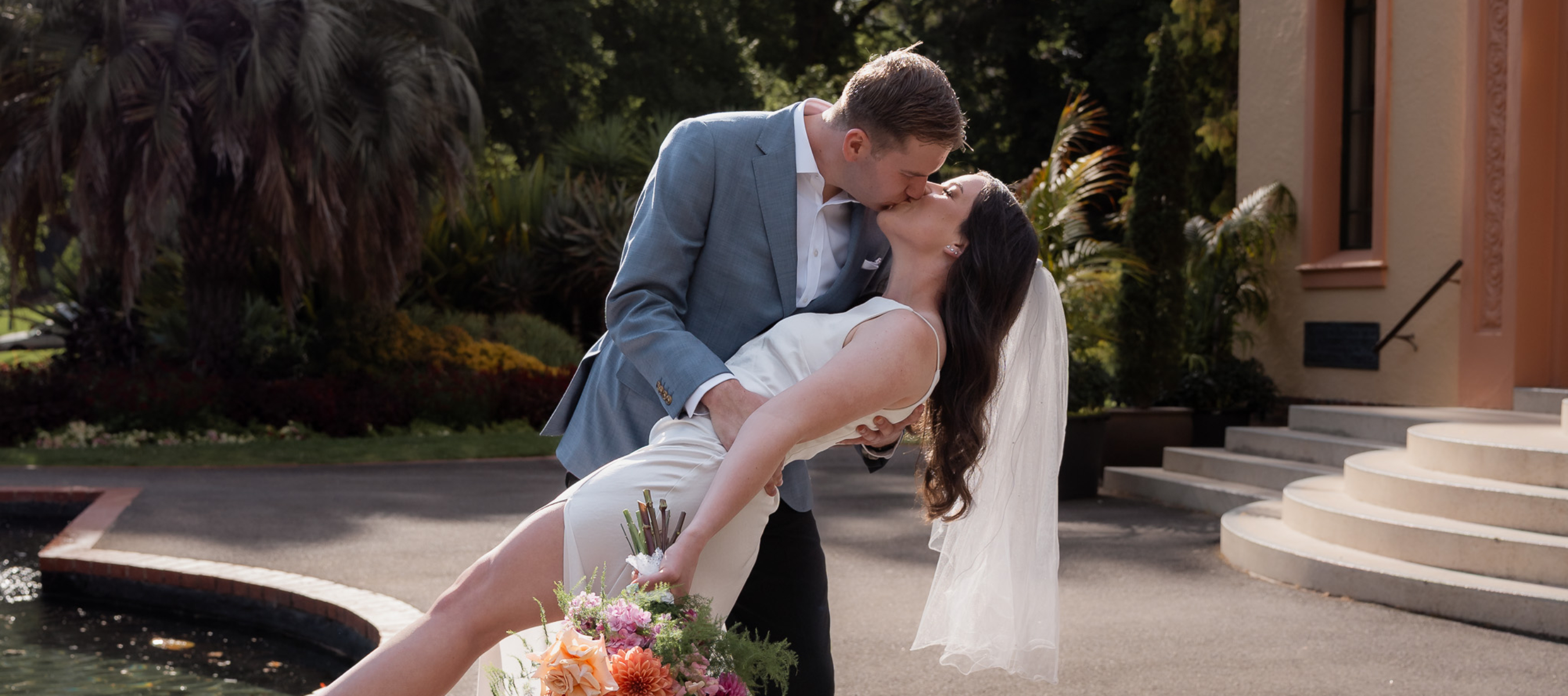 Bride and Groom dip kiss in the sunlight. Gardens in the background, a water foundtain and building with steps.