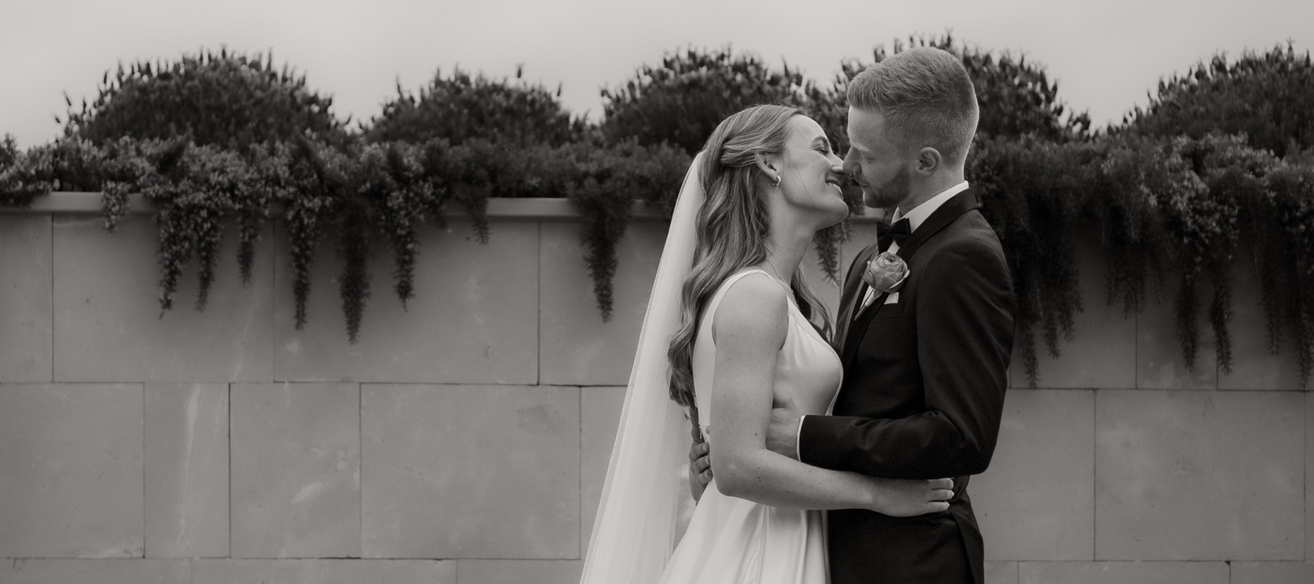 Bride and Groom close together, almost kissing and smiling. Stone wall in the back with garden bed. Black and white image.