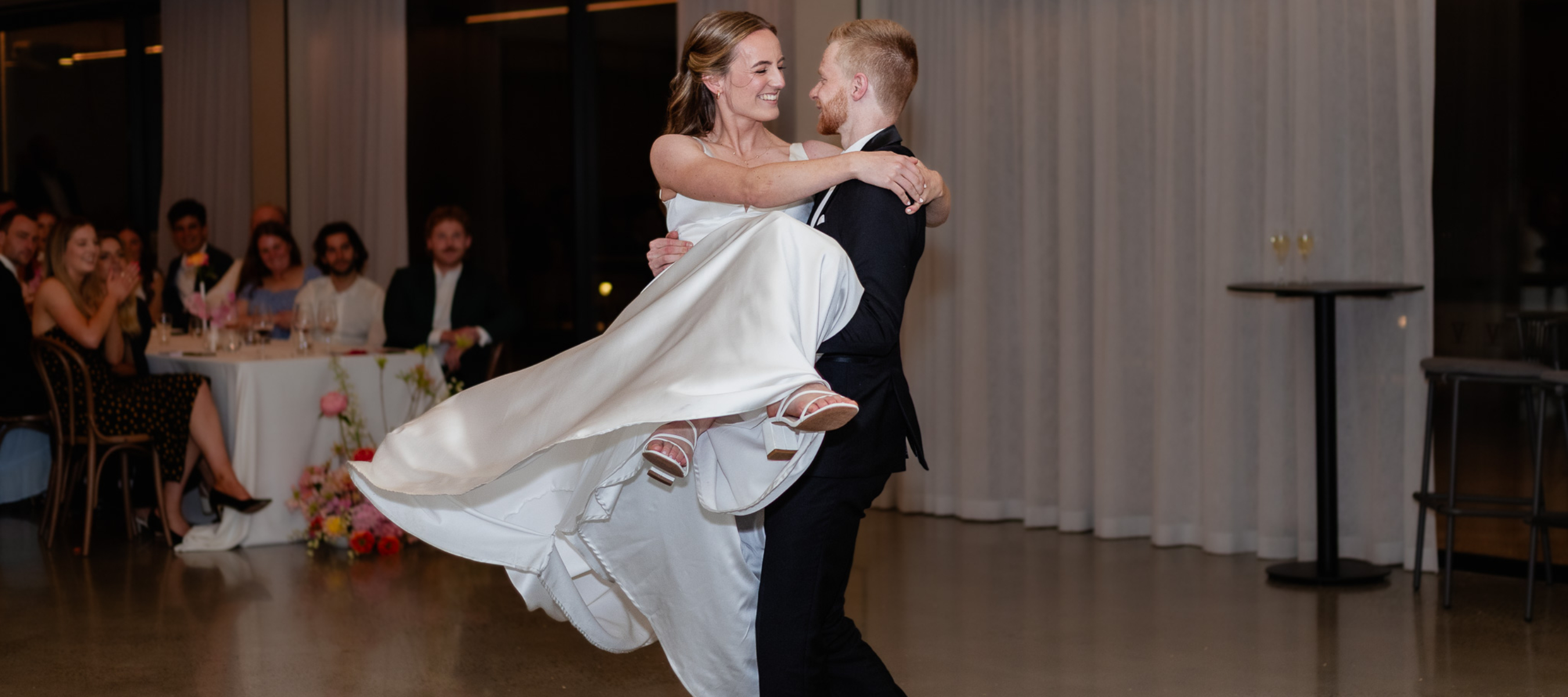 First dance photo of a Bride and Groom. Groom is spinning his bride. Guests in the background clapping and watching them dance.