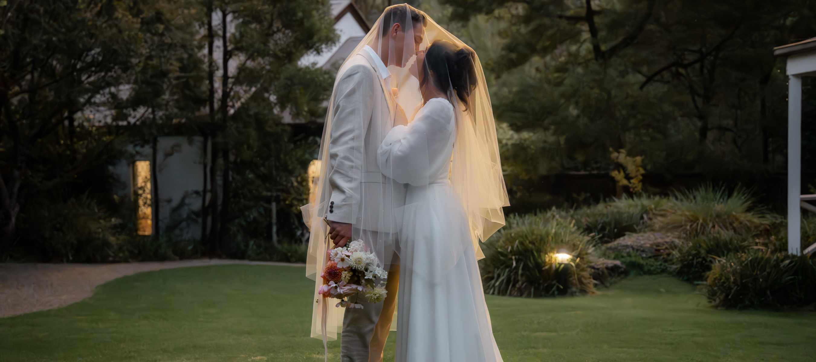 Bride and Groom under a wedding veil on grass in the sunset, chapel in the background